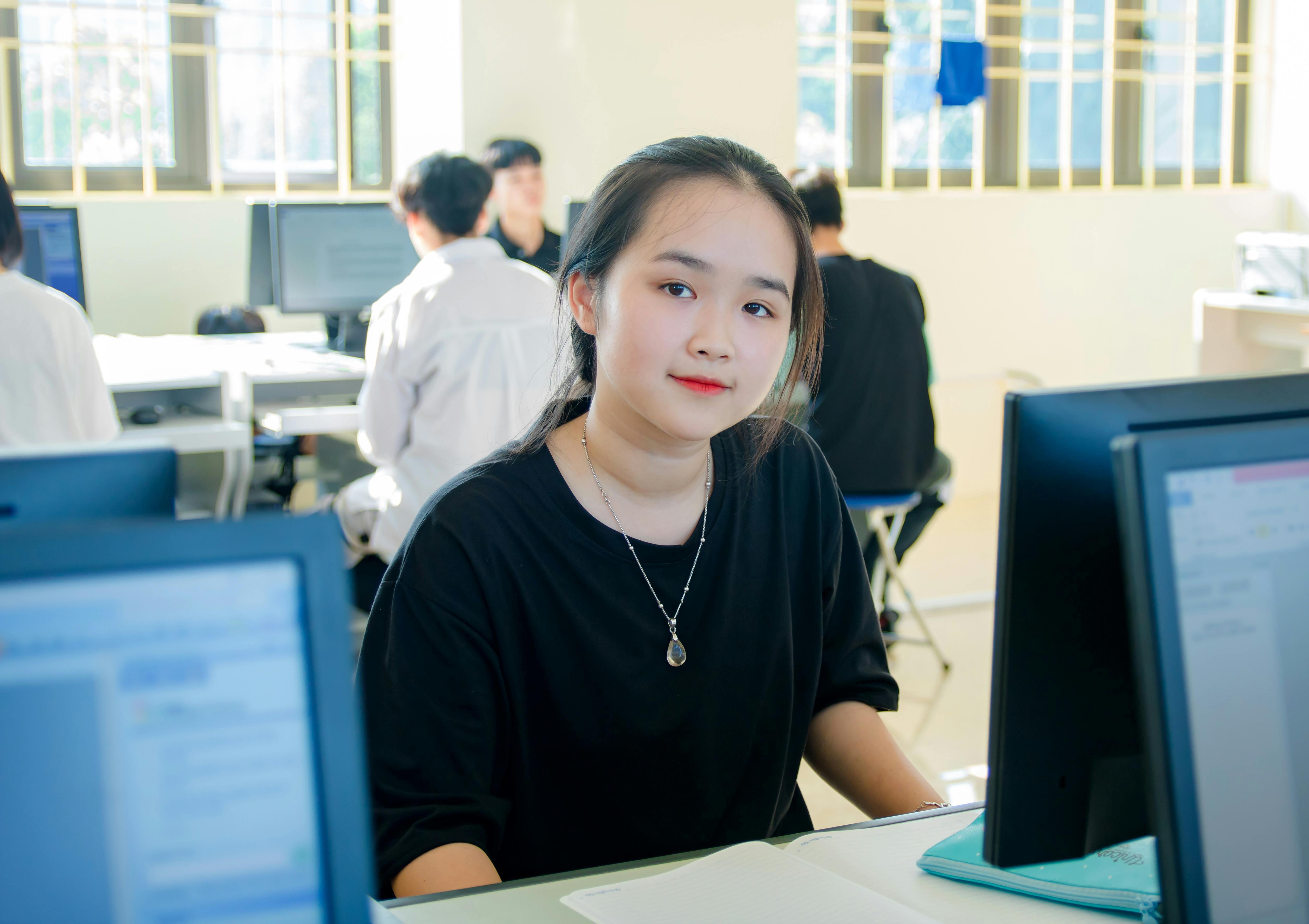 Student working on a laptop in a classroom or computer lab, seated at a desk with other students using devices in the background.
