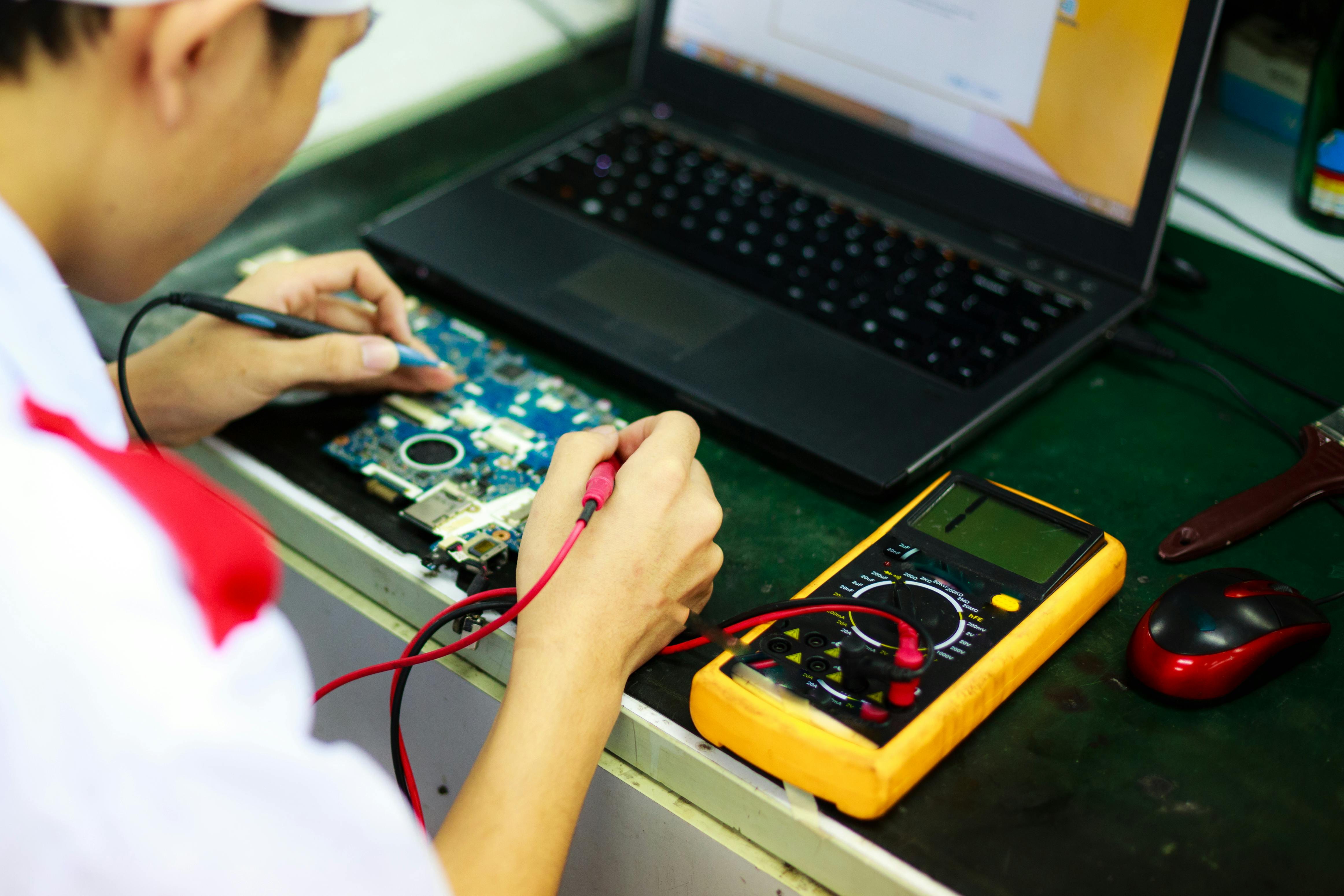 Technician testing a laptop motherboard with a multimeter to speed K–12 device repair turnaround time and reduce the need for loaner devices.